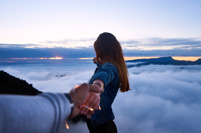 A "follow me" photo of a man holding hands with a brunette woman wearing a denim jacket on the top of the clouds